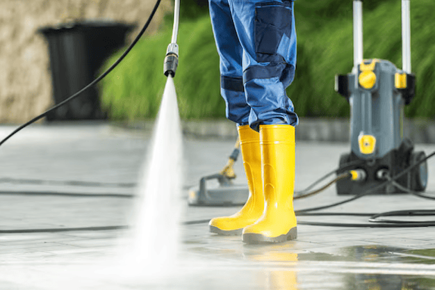 Worker cleaning surfaces with pressure washer while wearing yellow boots.